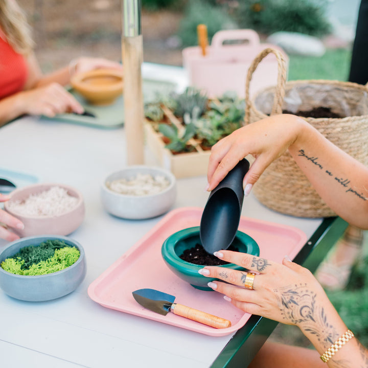 Person sitting at a table with gardening tools and plants, outdoors.